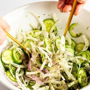 Hands mixing a salad with cucumbers, red onions, and herbs in a white bowl using two gold utensils.