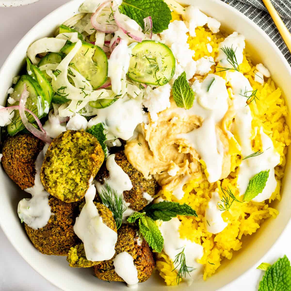 A bowl containing yellow rice, falafel, sliced cucumber, red onions, dill, feta cheese, and mint leaves, topped with hummus and drizzled with a white sauce.