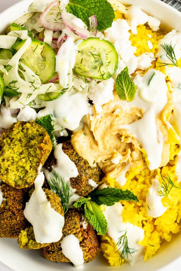 A bowl containing yellow rice, falafel, sliced cucumber, red onions, dill, feta cheese, and mint leaves, topped with hummus and drizzled with a white sauce.