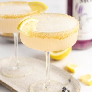 Two coupe glasses with yellow cocktails garnished with lemon slices and sugar rims on a beige tray. Purple-hued bottle in the background. Lavender sprigs and lemon pieces scattered around.