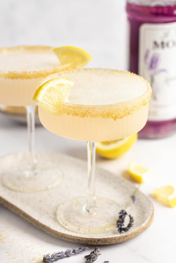 Two coupe glasses with yellow cocktails garnished with lemon slices and sugar rims on a beige tray. Purple-hued bottle in the background. Lavender sprigs and lemon pieces scattered around.