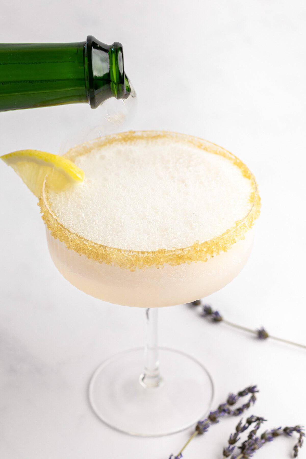 A green bottle pours liquid into a frothy glass with a sugared rim, garnished with a lemon slice. Lavender sprigs lie next to the glass on a white background.