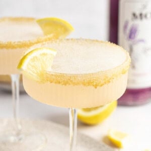 Two cocktails with frothy tops in coup glasses, each garnished with a lemon wedge and a sugar-rimmed edge. A bottle of purple liquid is partially visible in the background.