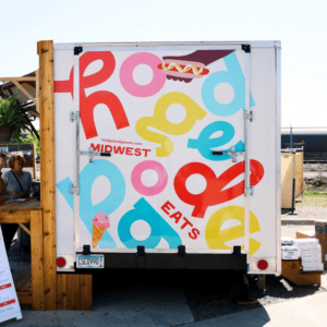 A food truck with a colorful sign that reads "Hodge Podge Eats Midwest" featuring playful letters, a hot dog, and an ice cream cone illustration. There are people and a menu sign next to the truck, drawing in fit foodie finds looking for their next delicious bite.