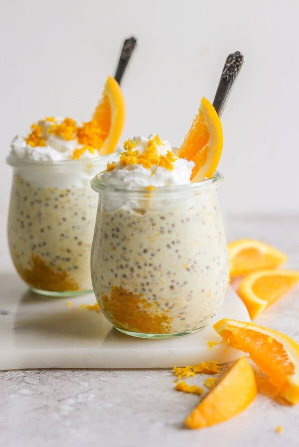 Two glass jars filled with creamy creamsicle overnight oats, topped with whipped cream and orange slices, arranged on a white surface with additional orange wedges nearby.