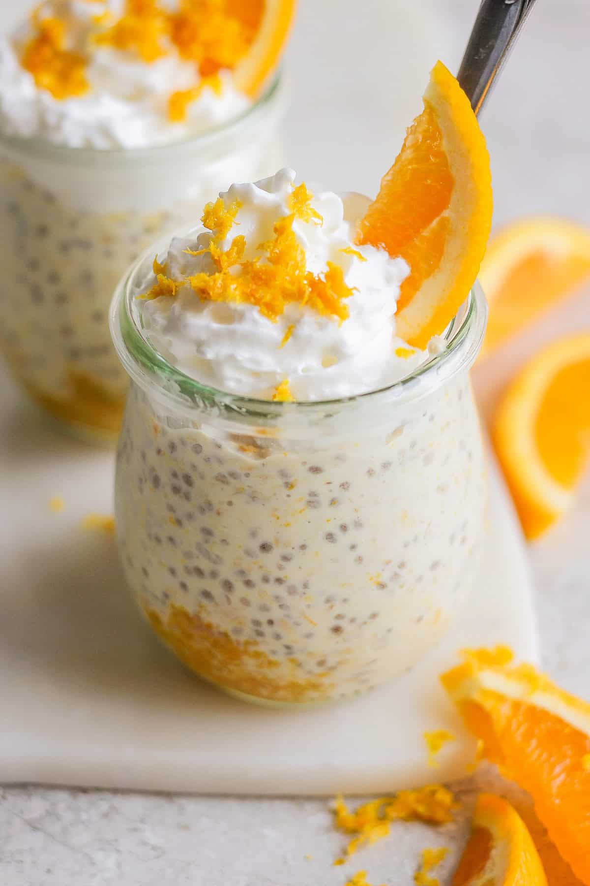 Two small glass jars filled with creamy chia seed pudding, reminiscent of creamsicle overnight oats, are topped with whipped cream, orange zest, and an orange slice. Orange slices on the side complete this delightful treat.