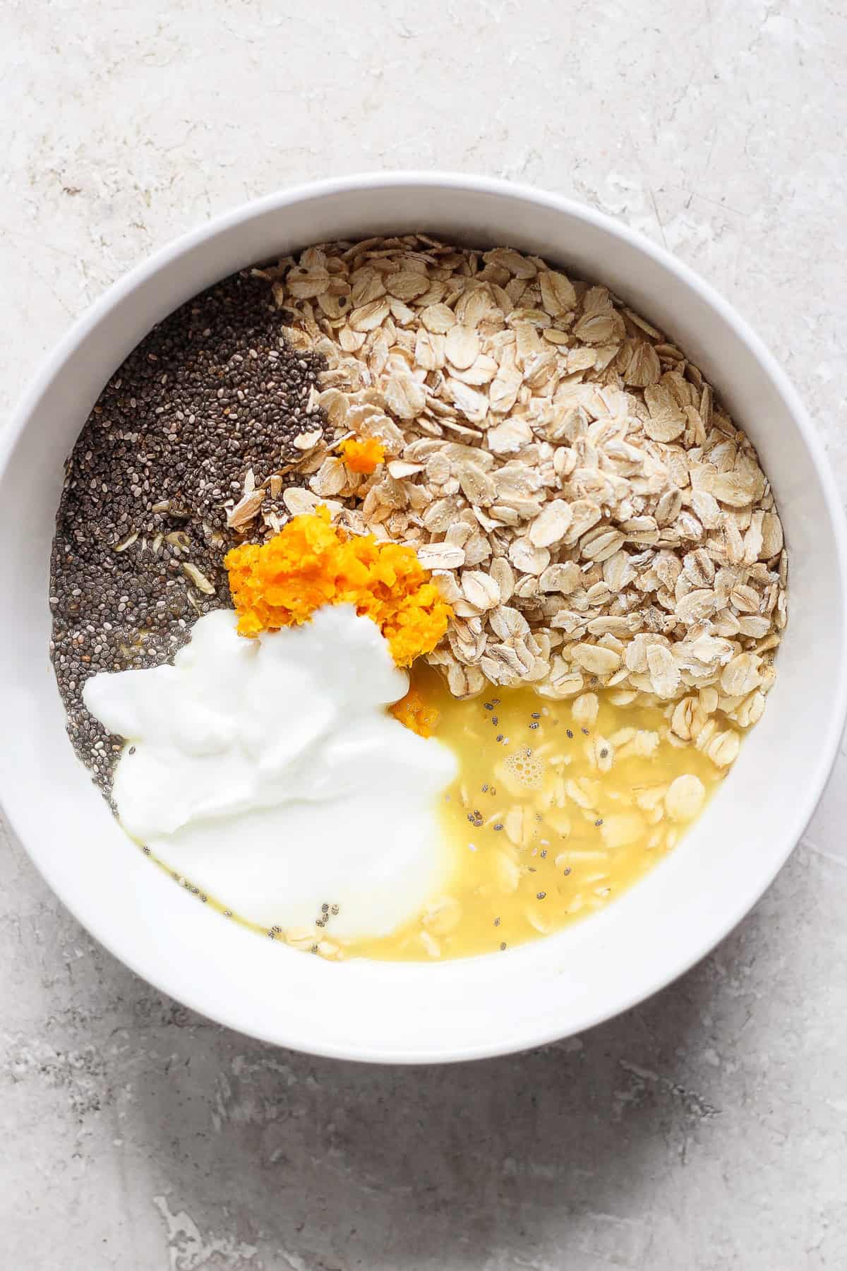 A white bowl contains oats, chia seeds, orange zest, yogurt, and liquid—likely milk or water—on a light-colored surface, creating a creamsicle overnight oats sensation.