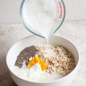 A hand pours milk from a measuring cup into a white bowl containing oats, chia seeds, orange zest, and yogurt, creating the perfect base for creamsicle overnight oats.