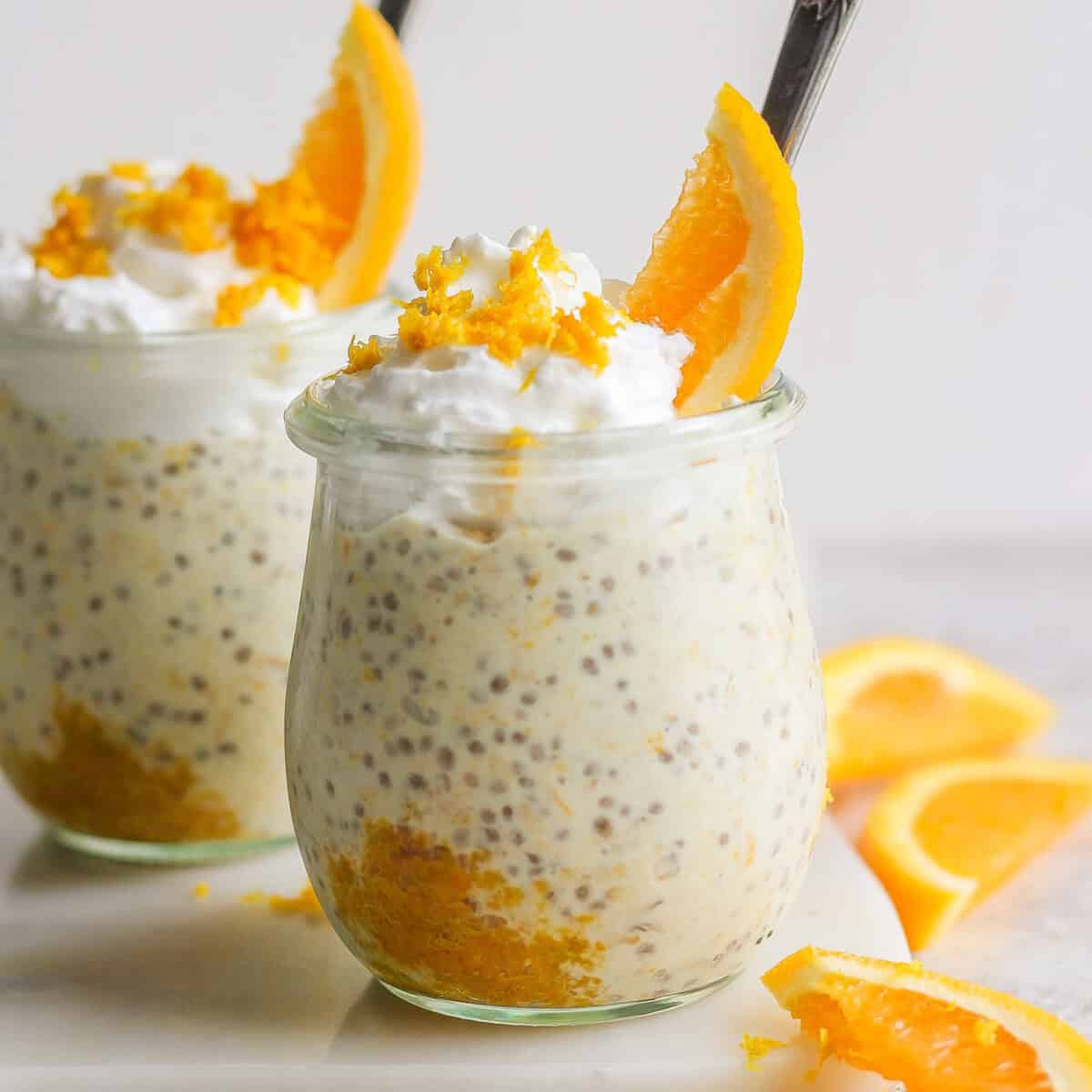 Two solid  jars filled with chia pudding topped with whipped cream, orangish  zest, and orangish  slices, each   with a spoon inserted. Slices of orangish  are placed beside the jars, giving disconnected  a vibes of creamsicle overnight oats.