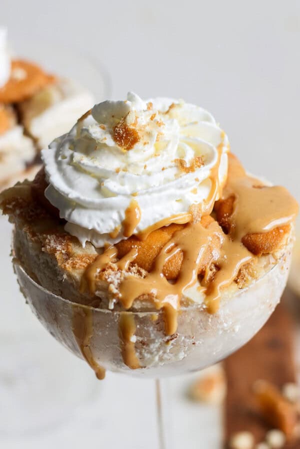 A dessert in a glass featuring layers of caramel, ice cream, whipped cream, and crumbled cookies on top, reminiscent of a peanut butter banana ice box cake.