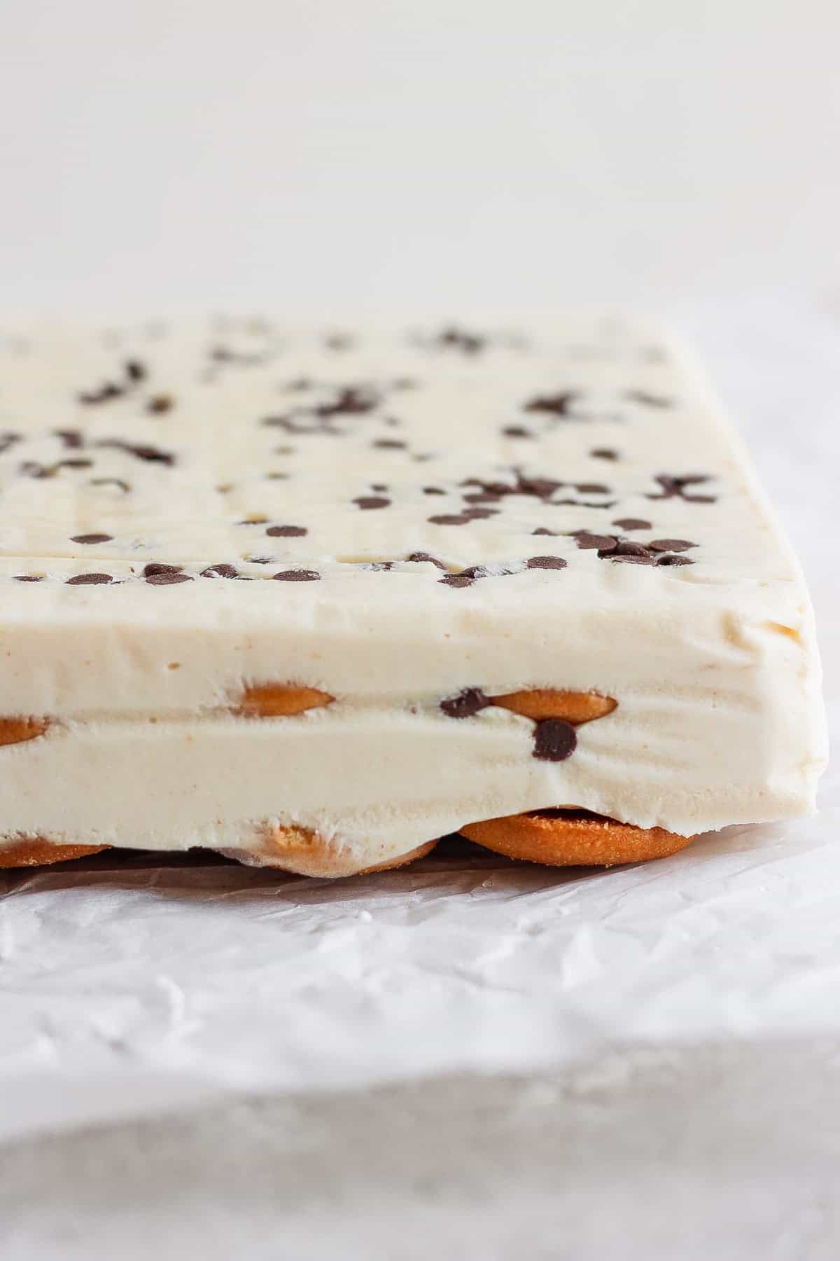 Close-up of a layered peanut butter banana ice box cake with a creamy white filling, chocolate chips on top, and a base of cookies, set on crinkled parchment paper.