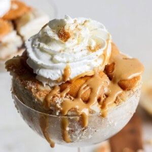 A dessert in a glass topped with whipped cream and caramel sauce. Crumbled biscuit pieces are visible, adding texture to the creamy base of this peanut butter banana ice box cake.