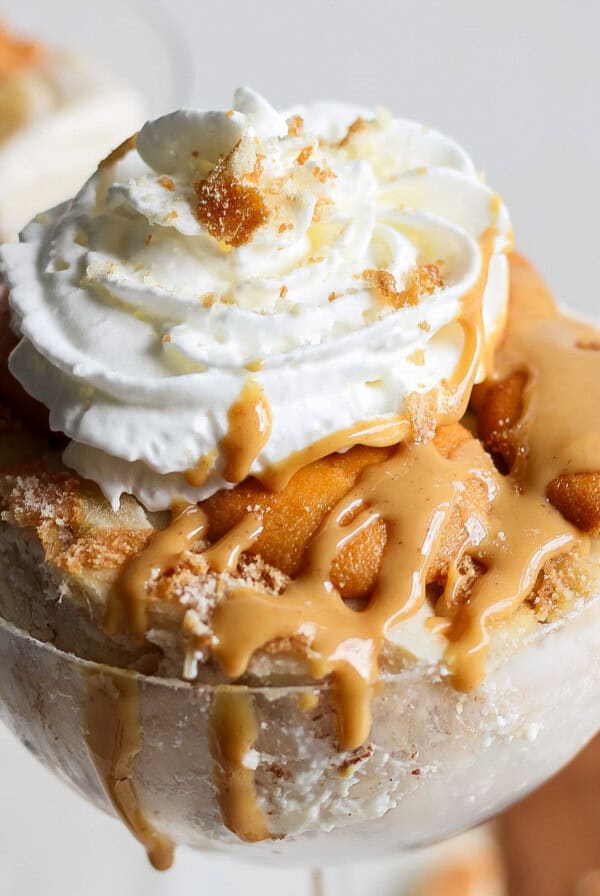 A dessert in a glass topped with whipped cream and caramel sauce. Crumbled biscuit pieces are visible, adding texture to the creamy base of this peanut butter banana ice box cake.