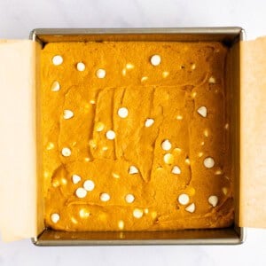 A square baking pan filled with unbaked batter topped with scattered white chocolate chips, lined with parchment paper on a marble countertop.