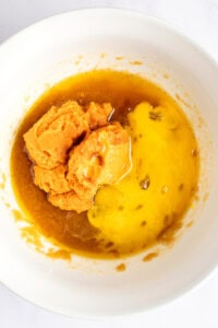 A mixing bowl contains mashed sweet potatoes, yolk-like liquid, and brown syrupy substance being prepared for a recipe.