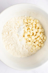 A white bowl containing a mixture of flour, baking powder, and white chocolate chips on a white background.