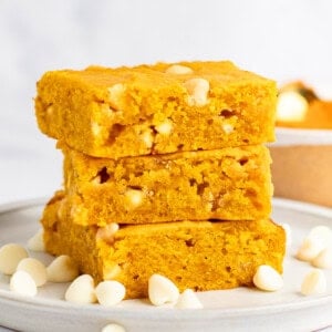 A stack of three pumpkin blondies with white chocolate chips on a white plate. Scattered white chocolate chips surround the stack.