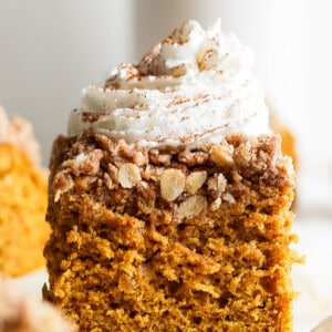 A slice of pumpkin coffee cake topped with whipped cream and a sprinkle of cinnamon on a white surface.