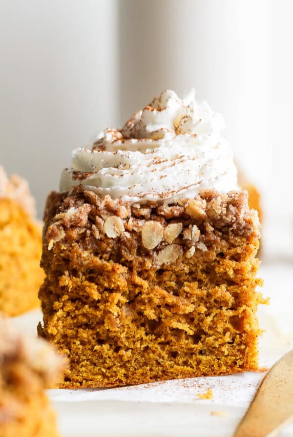 A slice of pumpkin coffee cake topped with whipped cream and a sprinkle of cinnamon on a white surface.