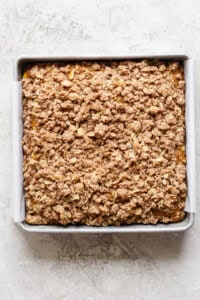 A square metal baking pan filled with a baked oatmeal fruit crumble topped with a crispy, golden-brown oat and cinnamon streusel. The pan is lined with parchment paper.