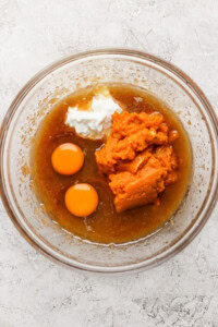 A clear glass bowl containing a mixture of ingredients including two raw eggs, pumpkin puree, white yogurt, and a brown liquid, is placed on a textured white surface.