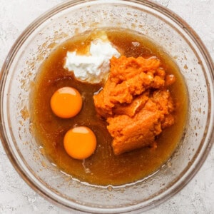 A clear glass bowl containing a mixture of ingredients including two raw eggs, pumpkin puree, white yogurt, and a brown liquid, is placed on a textured white surface.