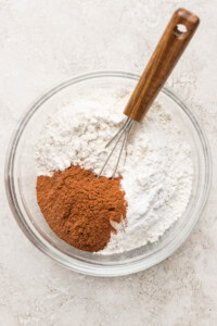 A clear glass bowl containing flour, cinnamon, and a small whisk with a wooden handle on a light-colored surface.