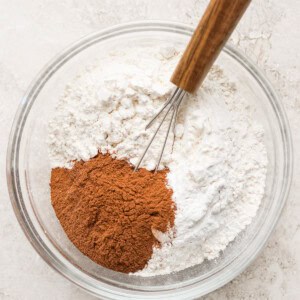 A clear glass bowl containing flour, cinnamon, and a small whisk with a wooden handle on a light-colored surface.