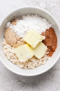 A white bowl containing oats, brown sugar, flour, cinnamon, and two slabs of butter on a light-colored surface.