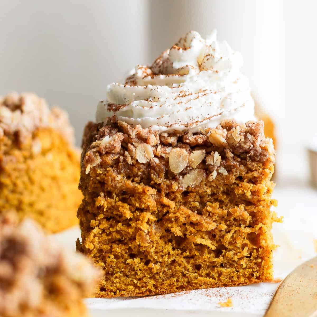 A slice of pumpkin crumb cake topped with whipped cream and a sprinkle of cinnamon, placed on a white surface.