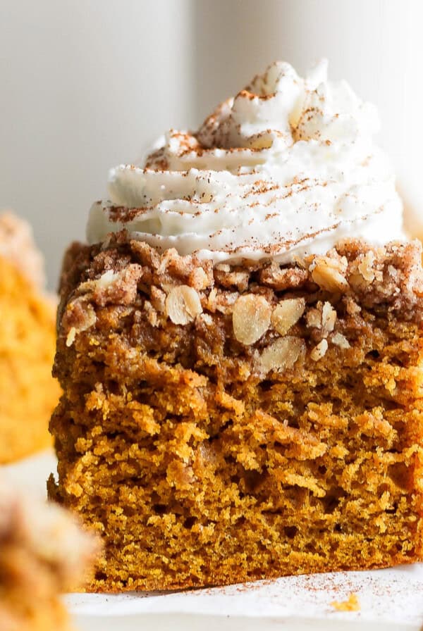 A slice of pumpkin crumb cake topped with whipped cream and a sprinkle of cinnamon, placed on a white surface.