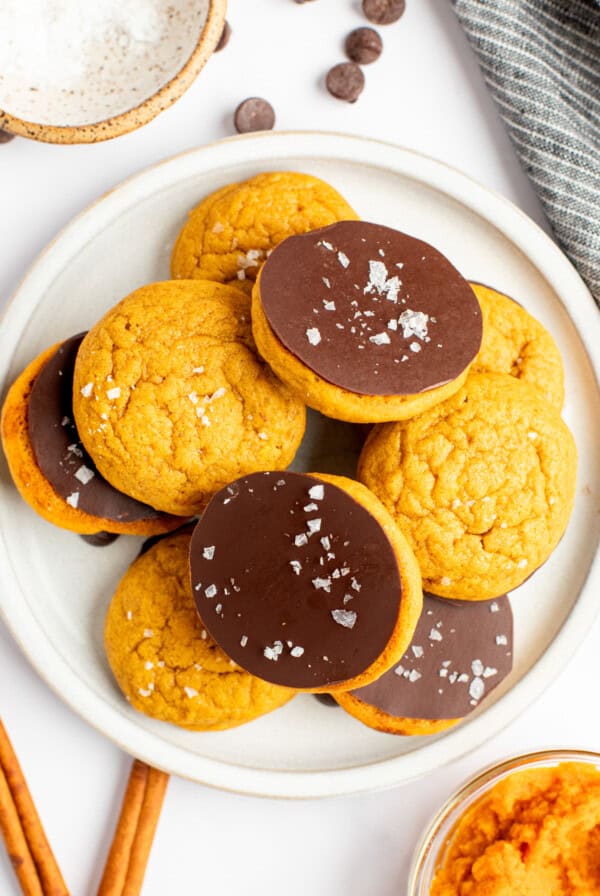 A white plate holds several cookies, half of which are chocolate-topped and sprinkled with coarse sea salt. The other half are plain. Nearby are a bowl of dip, a cloth, chocolate chips and cinnamon sticks.