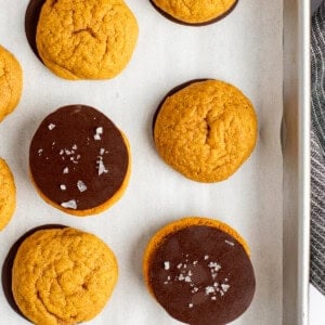 A baking tray with parchment paper holds nine round orange cookies, some plain and some half-dipped in chocolate, with a few sprinkled with sea salt.