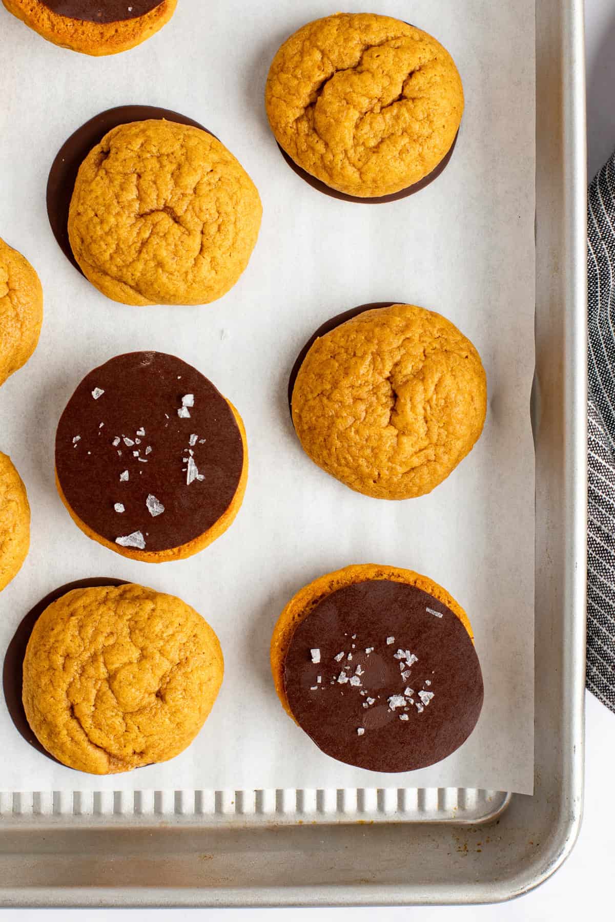 A baking tray with parchment paper holds nine round orange cookies, some plain and some half-dipped in chocolate, with a few sprinkled with sea salt.