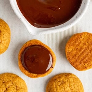 Cookies being dipped in melted chocolate from a white bowl, with additional cookies placed nearby on a white surface.