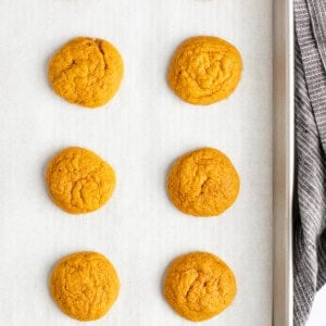 Eight freshly baked cookies on a parchment-lined baking sheet with a gray cloth visible on the right side.