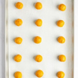 A baking tray with 15 evenly spaced, round, orange dough balls placed on a parchment-lined surface.