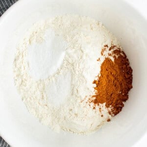 A white bowl containing flour, cinnamon, and baking powder, with a striped cloth partially visible in the background.