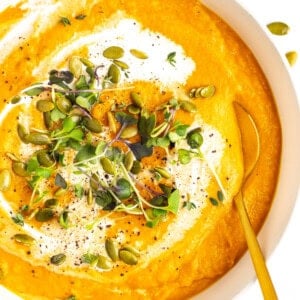 A bowl of creamy orange soup garnished with fresh microgreens, pumpkin seeds, black pepper, and a swirl of cream. A golden spoon rests inside the bowl.