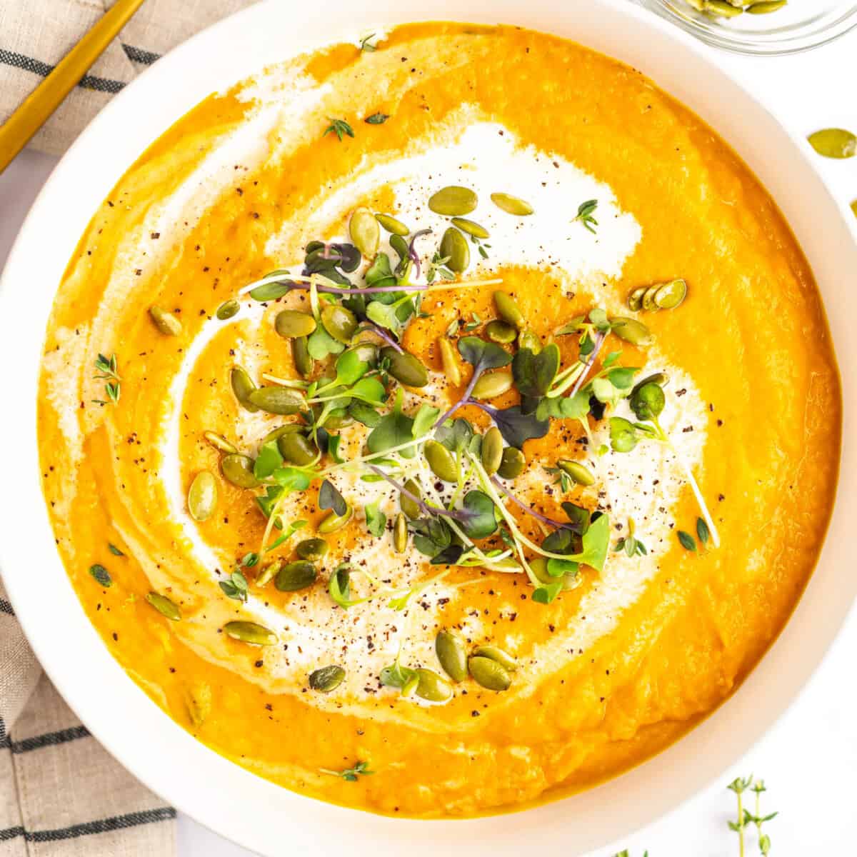 A bowl of creamy orange soup topped with microgreens, pumpkin seeds, and a drizzle of cream. Black pepper and a sprig of thyme garnish the soup. A striped napkin is partially visible on the side.