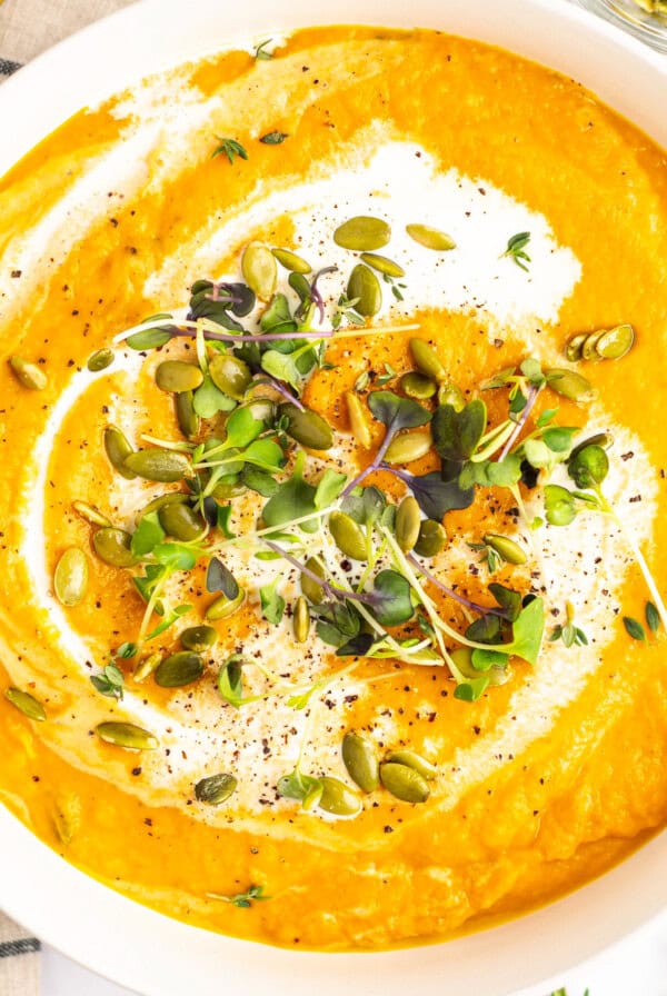 A bowl of creamy orange soup topped with microgreens, pumpkin seeds, and a drizzle of cream. Black pepper and a sprig of thyme garnish the soup. A striped napkin is partially visible on the side.