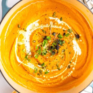 A pot of creamy carrot soup garnished with a swirl of cream, microgreens, pepitas, and black pepper, alongside a piece of bread and herbs.