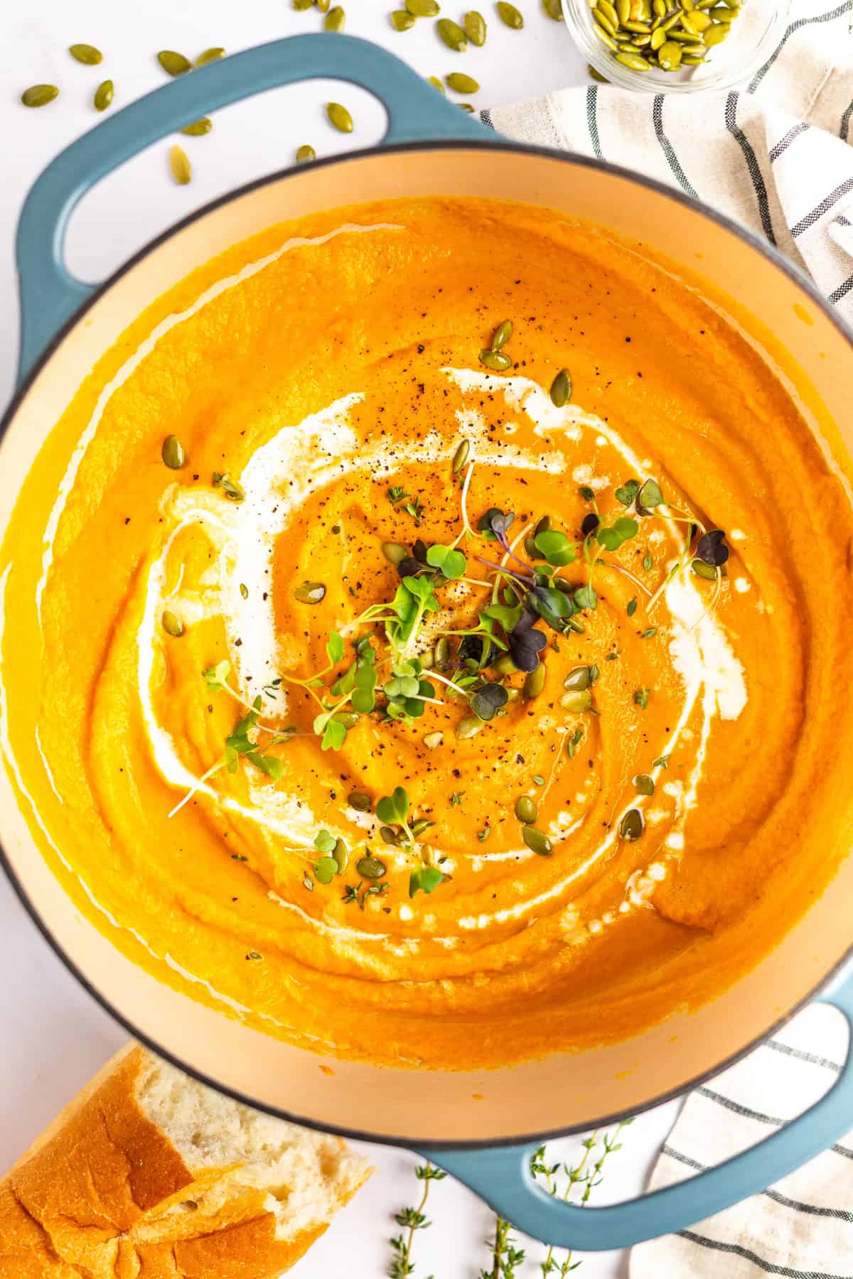 A pot of creamy carrot soup garnished with a swirl of cream, microgreens, pepitas, and black pepper, alongside a piece of bread and herbs.