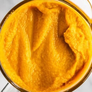 A top view of a glass container filled with smooth, bright orange pumpkin puree on a white marble surface.