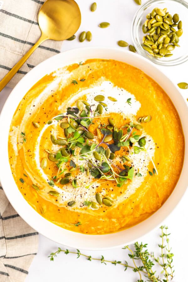 A bowl of creamy orange soup garnished with microgreens, pumpkin seeds, and a swirl of cream. A gold spoon, sprigs of fresh herbs, and a small bowl of pumpkin seeds are placed nearby.