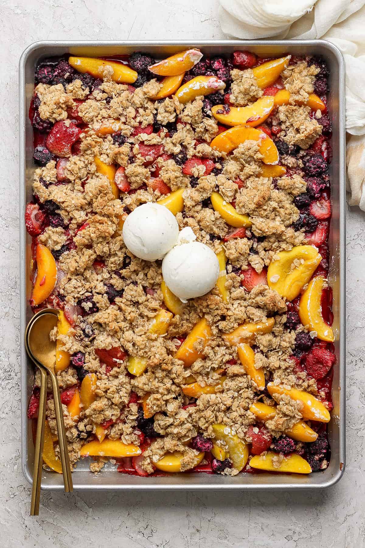 A rectangular baking tray filled with a mixed berry and peach crumble topped with two scoops of vanilla ice cream. A gold spoon rests on the left side of the tray.