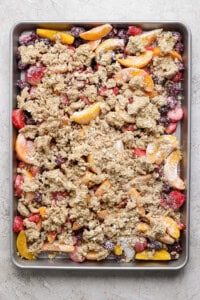 A baking tray filled with a mixture of sliced fruits, including peaches and berries, topped with a crumbly oat mixture, ready to be baked.