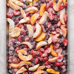 A baking sheet filled with an assortment of frozen fruit slices including peaches, strawberries, and blackberries arranged evenly across the surface.