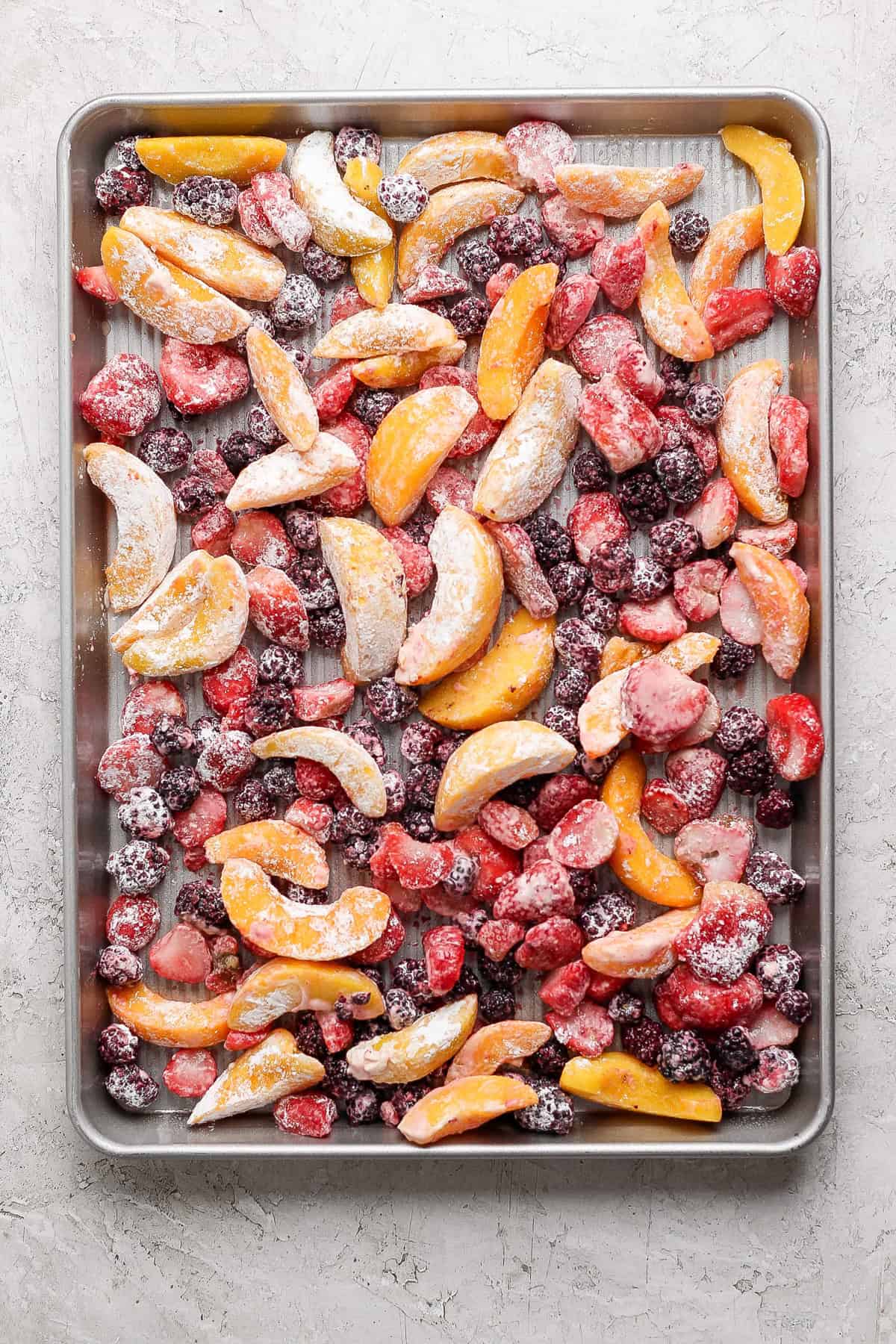 A baking sheet filled with an assortment of frozen fruit slices including peaches, strawberries, and blackberries arranged evenly across the surface.