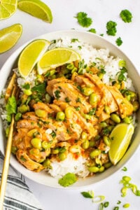 A bowl of white rice topped with sliced chicken, soybeans, and a creamy sauce, garnished with lime wedges and cilantro. A pair of gold chopsticks rests beside the bowl. Lime slices are scattered around.